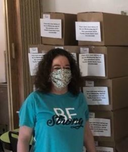 A woman wearing a protective face mask stands in front of stacks of boxed meals